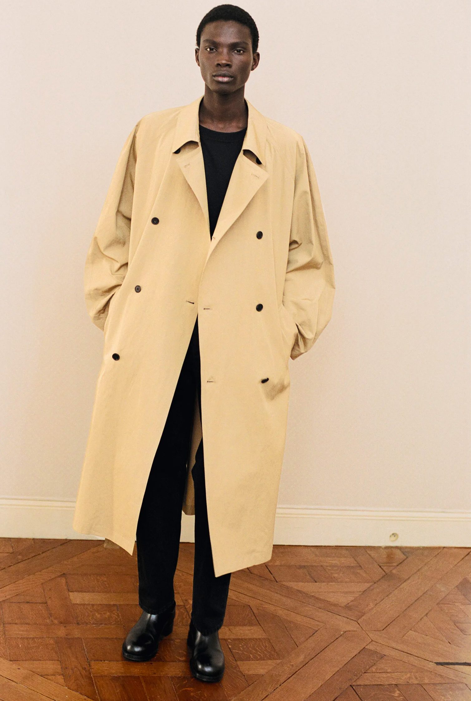 Total look The Row: il modello indossa un lungo cappotto color kaki sopra un completo monocromatico nero e stivali in pelle nera