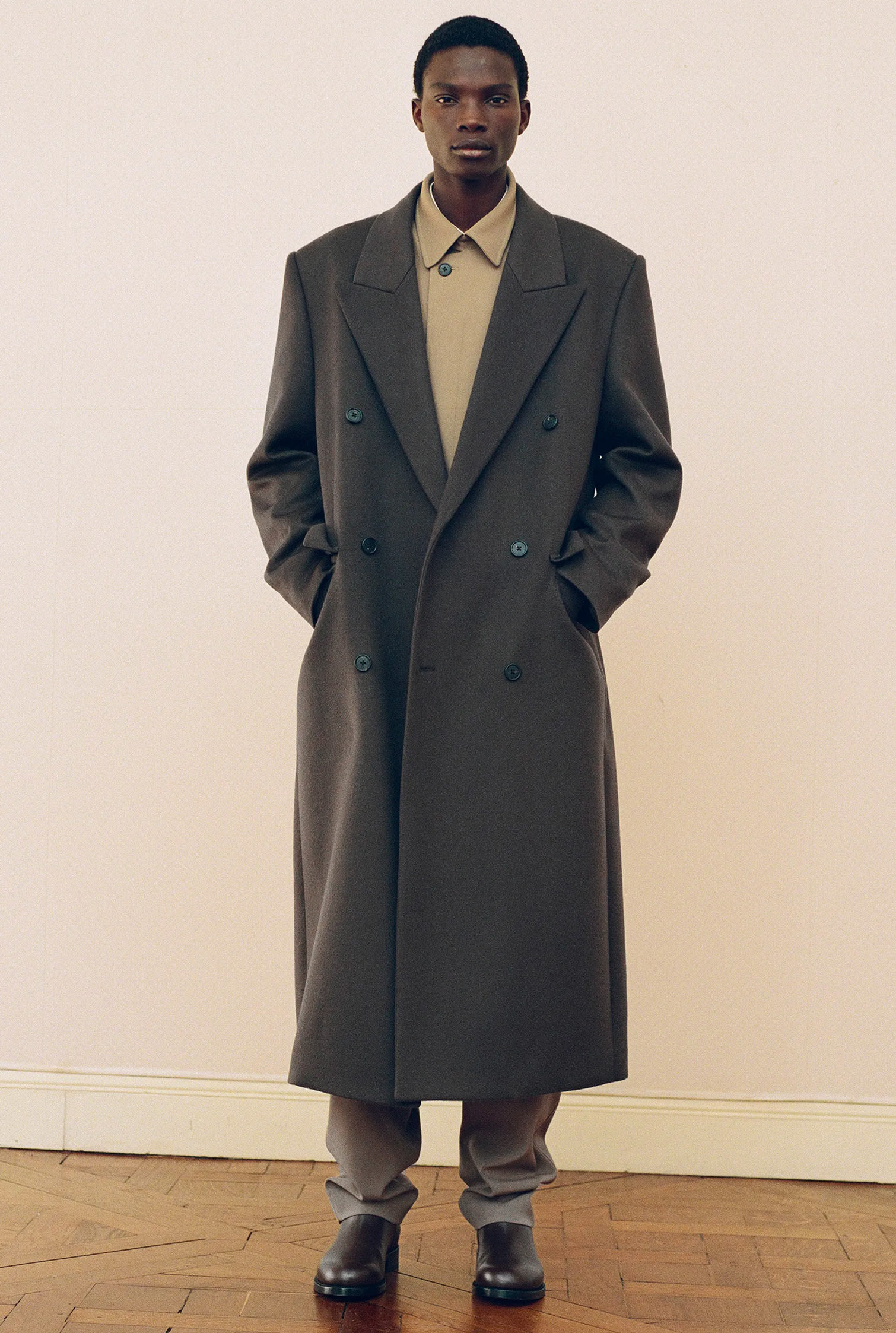 Il modello indossa un total look The Row composto da cappotto lungo in cashmere verde sopra una giacca di lana kaki con pantaloni kaki e stivali neri